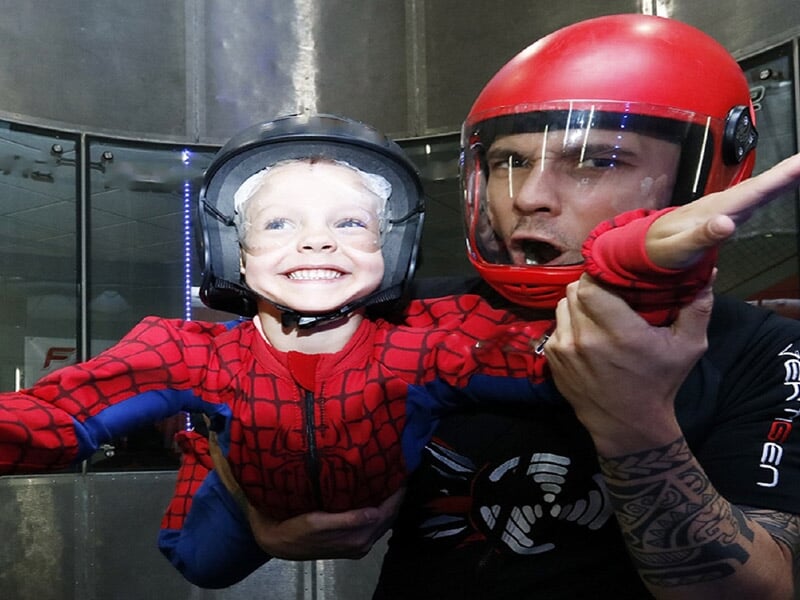 Enfant souriant en costume rouge et bleu, guidé par un instructeur casque rouge dans un simulateur de vol.