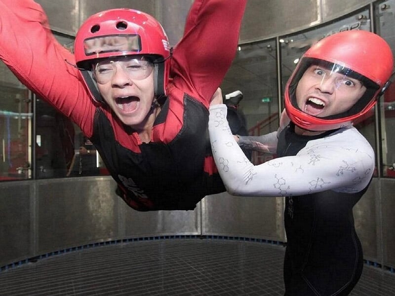 Femme criant d’excitation, tenue par un instructeur dans un tunnel de vent, casque rouge et lunettes de protection.