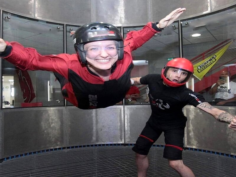 Femme souriante en chute libre indoor, guidée par un instructeur en casque rouge.