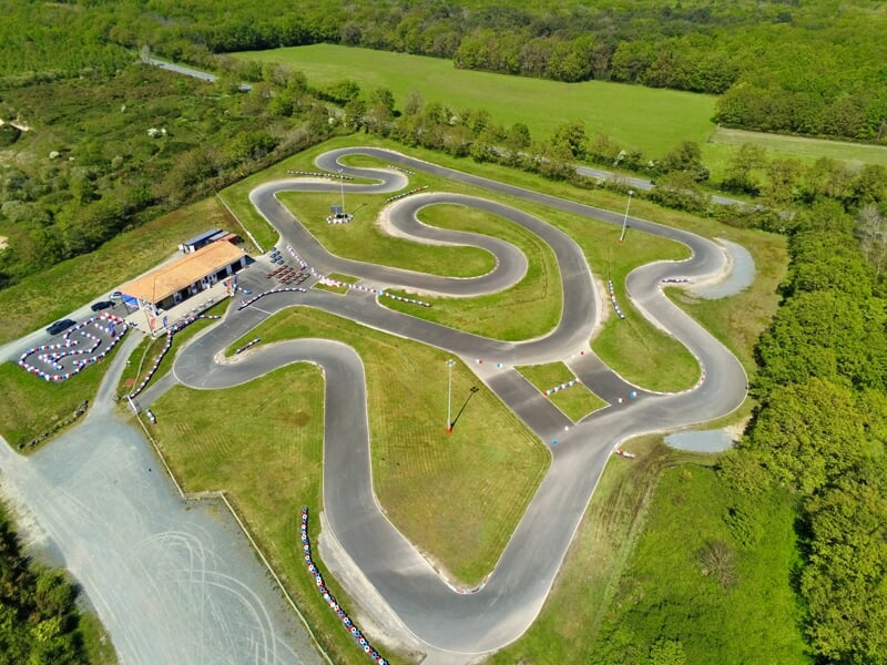 Vue aérienne d’un circuit de karting sinueux entouré de verdure. Bâtiment d’accueil avec parking visible à l’entrée. Piste asphaltée avec virages serrés et barrières de sécurité colorées.