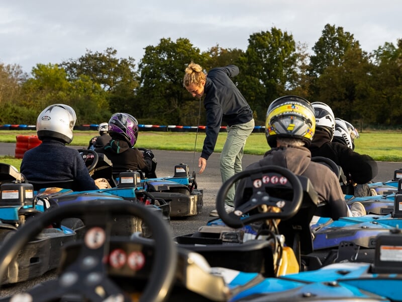 Briefing avant course sur un circuit de karting. Un instructeur s'adresse aux pilotes assis dans leurs karts bleus, tous équipés de casques. Lumière dorée du matin et arbres en arrière-plan.