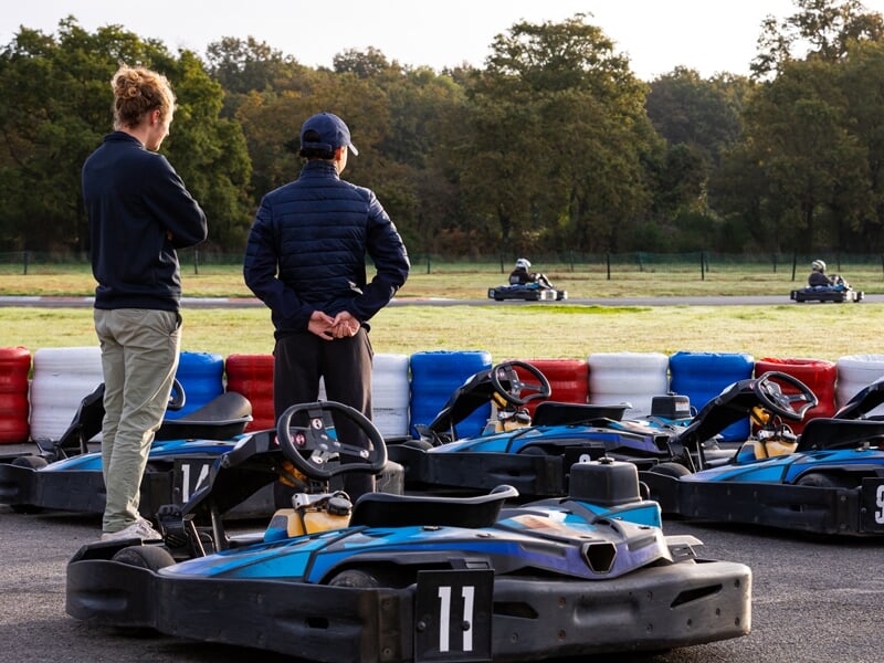 Karting en plein air avec plusieurs karts bleus alignés près des stands. Deux hommes observent la course en arrière-plan, l'un les bras croisés et l'autre avec les mains dans le dos. Barrières de sécu