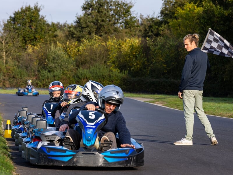 Départ d’une course de karting avec des pilotes casqués dans des karts bleus alignés sur la piste. Un homme en survêtement clair tient un drapeau à damier en surveillant les participants. Environnemen