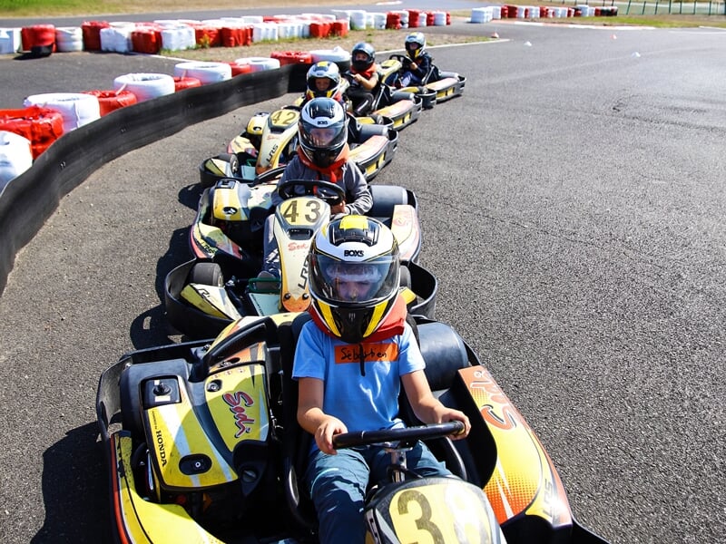 Enfants en tenue de course et casques intégraux prêts à démarrer une session de karting. Karts jaunes numérotés alignés sur la piste avec des protections rouges et blanches autour du circuit.
