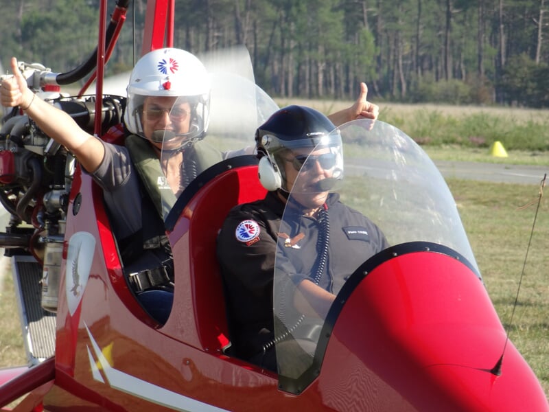 Deux personnes dans un ULM rouge, prêtes à décoller, souriantes et faisant un pouce levé sur un terrain d'aviation.