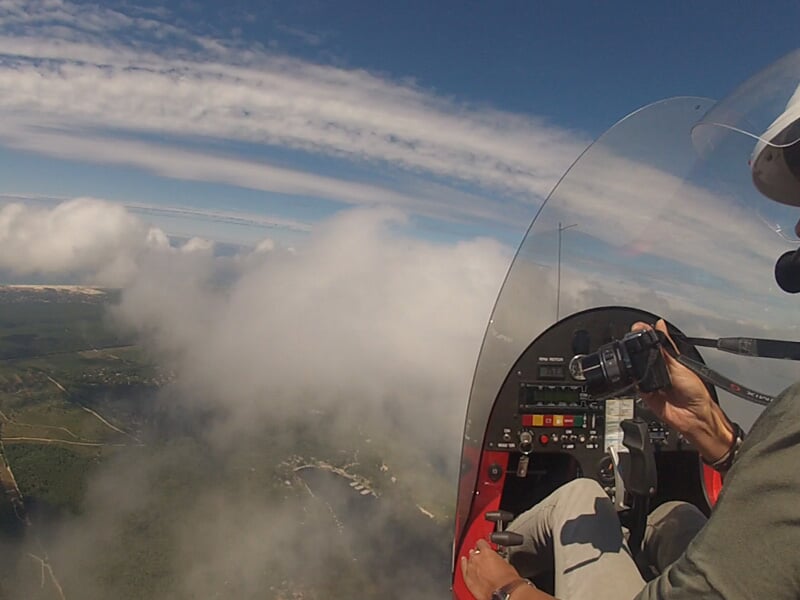 Vue depuis l'intérieur d'un ULM en vol, avec un pilote aux commandes et un paysage nuageux en dessous.