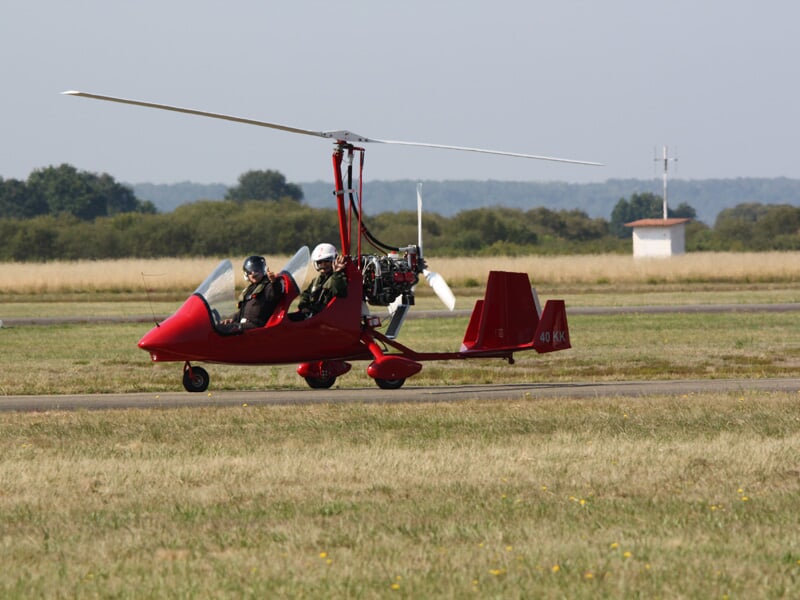 ULM rouge au sol sur une piste d'herbe, avec deux personnes à bord portant des casques de pilote.