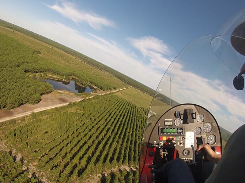 Vue en vol d'un ULM survolant des champs verts et une rivière sous un ciel bleu avec quelques nuages.