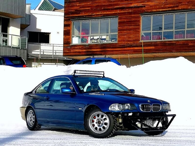 BMW bleue équipée pour circuit sur glace, avec pare-chocs renforcé, roulant sur neige devant un bâtiment moderne.