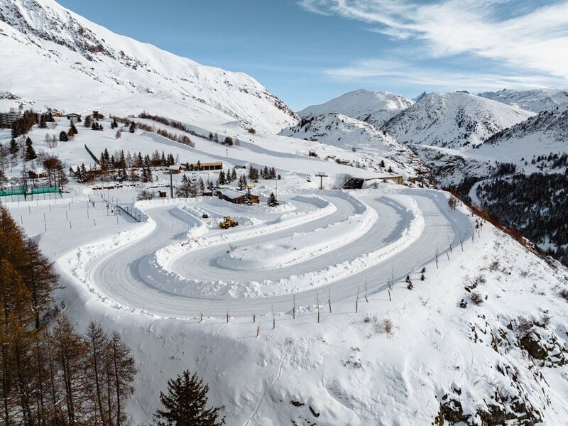Vue aérienne d'un circuit de glace sinueux en montagne enneigée, avec des arbres et des bâtiments autour.