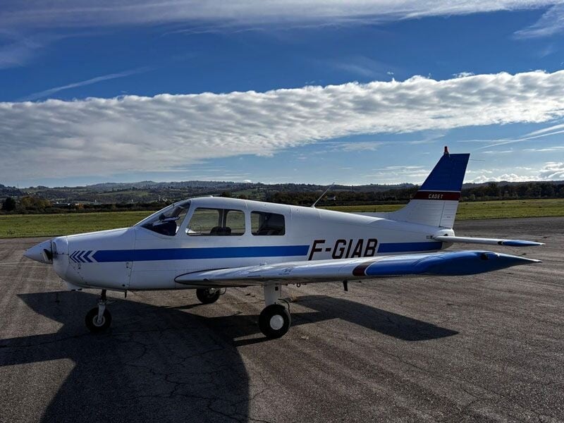 Petit avion blanc et bleu stationné sur une piste d'aéroport sous un ciel partiellement nuageux.