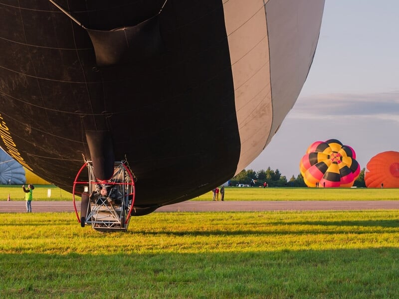 Gros plan sur la base d'une montgolfière noire posée sur un champ avec d'autres ballons colorés en arrière-plan.