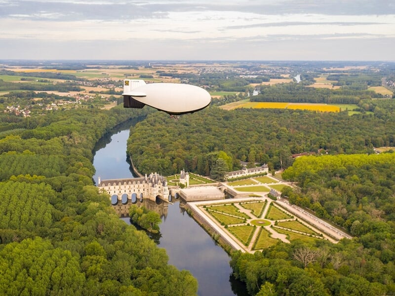 Vue aérienne d'un dirigeable blanc volant au-dessus d'un château entouré de forêts et d'un cours d'eau.