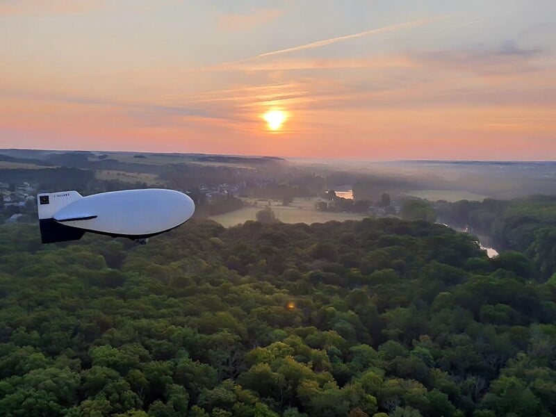 Dirigeable blanc survolant une forêt dense au coucher du soleil avec un ciel aux teintes orangées.