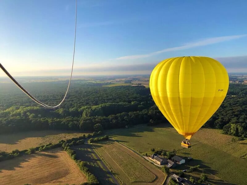 Montgolfière jaune volant au-dessus de champs et forêts sous un ciel clair au lever ou coucher du soleil.
