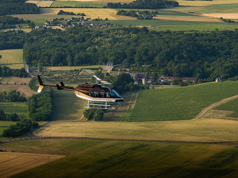 Hélicoptère survolant une campagne verdoyante avec un château en arrière-plan.