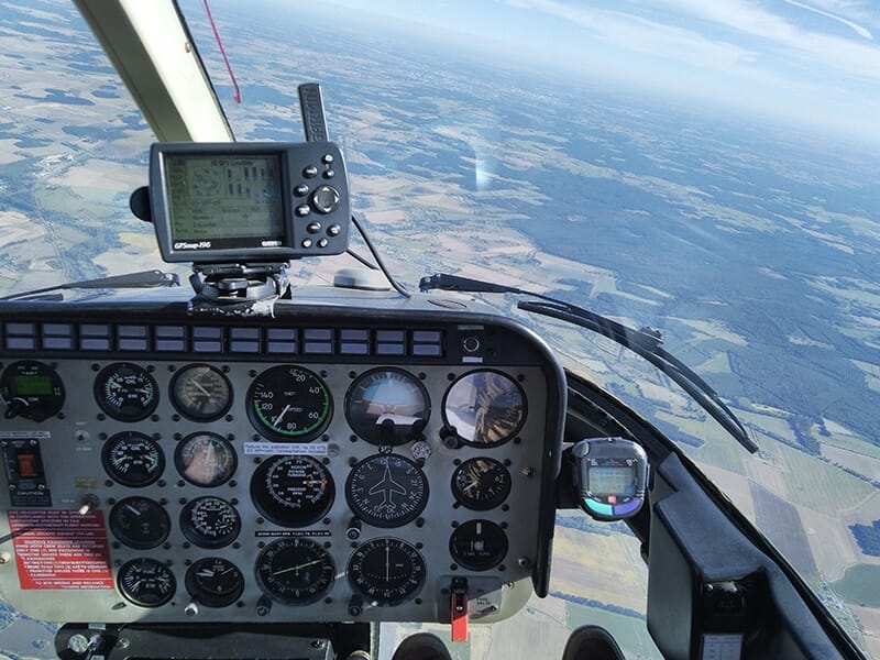 Vue depuis le cockpit d’un hélicoptère en vol, avec les instruments de bord et un paysage rural à l’horizon.