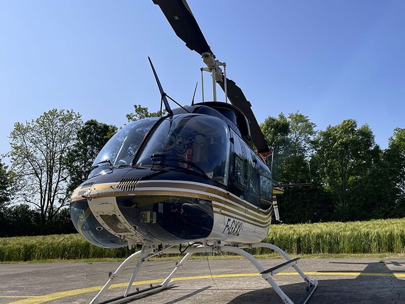 Hélicoptère posé sur un héliport entouré d’arbres et de champs sous un ciel bleu.