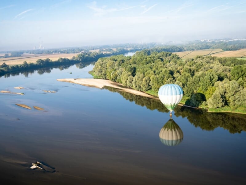 Vue aérienne d'une montgolfière rayée en bleu et blanc flottant au-dessus d'une rivière sinueuse. Le reflet de la montgolfière se mire dans l'eau claire. La scène est bordée par des forêts denses et d
