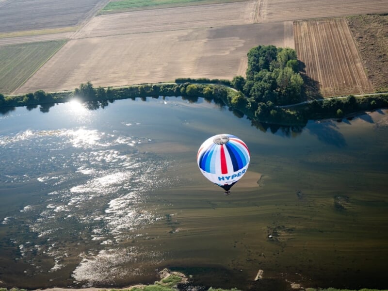 Montgolfière multicolore avec des bandes bleues, blanches et rouges survolant un plan d'eau, avec un reflet lumineux du soleil, entouré de vastes champs agricoles et d'une petite île boisée. L'image c
