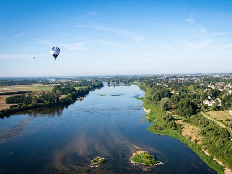 Montgolfière colorée survolant un large cours d'eau bordé de verdure luxuriante et de zones résidentielles. La rivière serpente à travers un paysage agricole avec des îlots de végétation. Le ciel clai