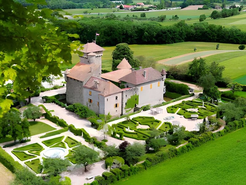 Château entouré d'un jardin à la française avec des haies taillées et une fontaine, en pleine campagne.