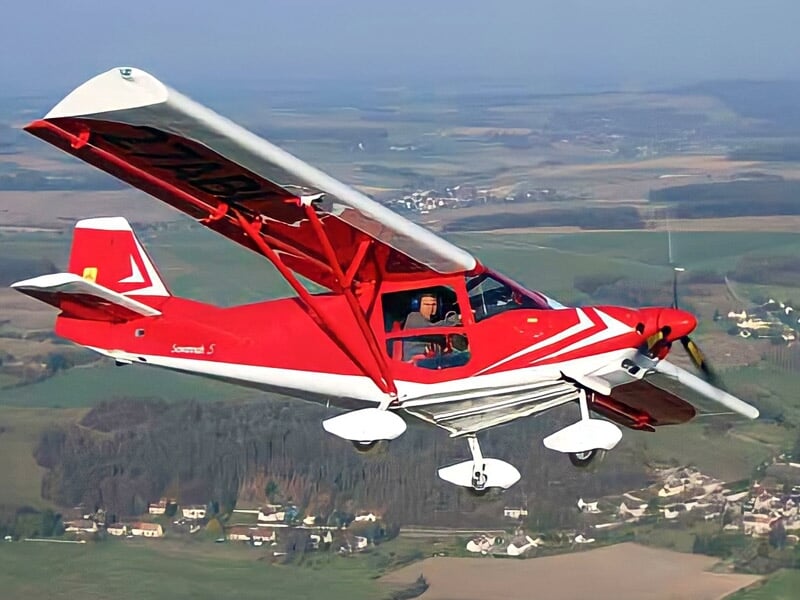 Petit avion rouge et blanc en vol au-dessus d'un paysage rural avec champs et forêts à l'horizon.