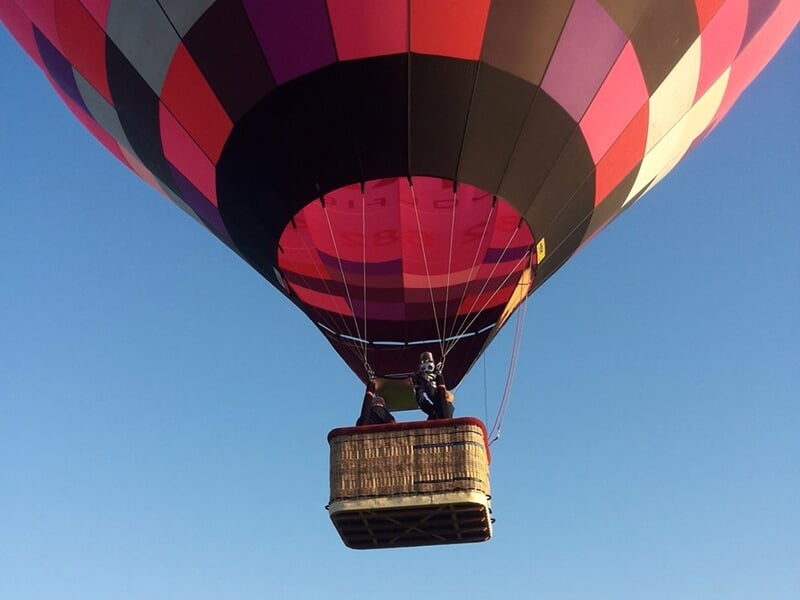 Montgolfière rose et violette en vol, avec une nacelle contenant deux passagers sous un ciel bleu clair