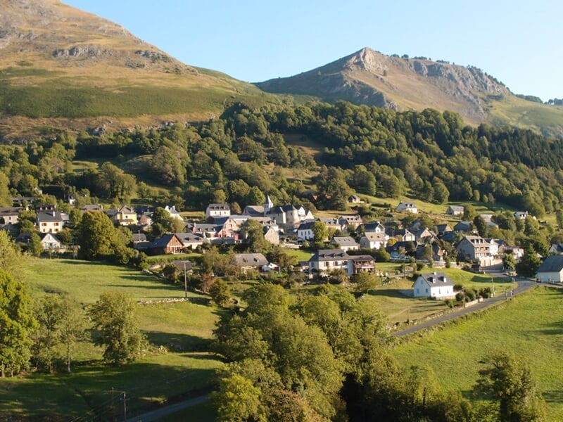 Village de montagne entouré de verdure, niché au pied de collines boisées et de sommets rocheux sous un ciel dégagé