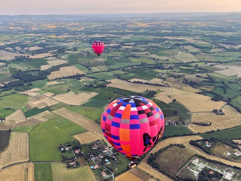 Deux montgolfières colorées survolant une campagne vallonnée, composée de champs cultivés et de hameaux