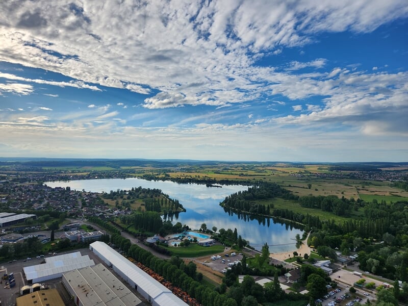 Vue aérienne d'un lac entouré de verdure et de bâtiments sous un ciel partiellement nuageux