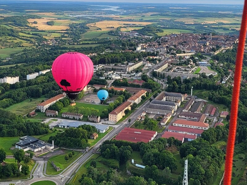 Vue aérienne de deux montgolfières, une rose et une bleue, flottant au-dessus de bâtiments entourés d'arbres, avec une ville en arrière-plan