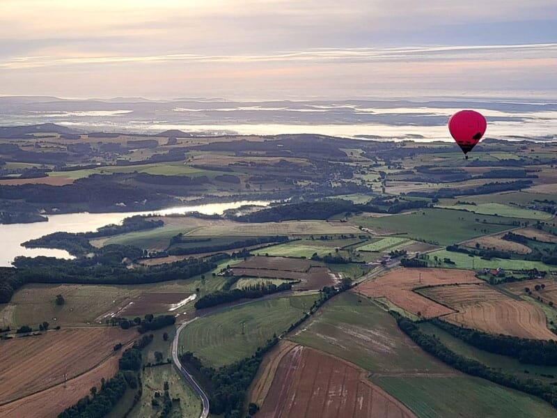 Montgolfière rose survolant une campagne vallonnée avec des champs et des cours d'eau, sous un ciel clair au lever du soleil