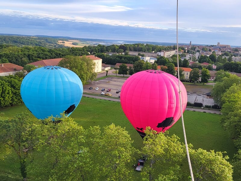 Montgolfières rose et bleue en vol bas au-dessus d'une pelouse, entourées d'arbres, avec la ville visible à l'arrière-plan