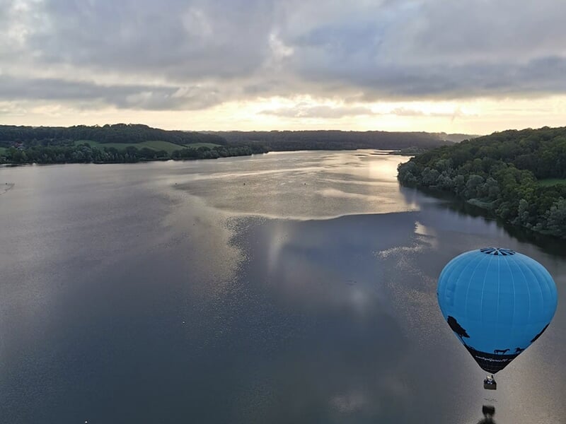 Montgolfière flottant au-dessus d'un grand lac entouré de collines boisées sous un ciel nuageux.