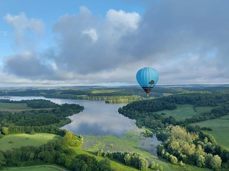 Montgolfière bleue survolant une région boisée et un lac paisible, avec une vue panoramique.