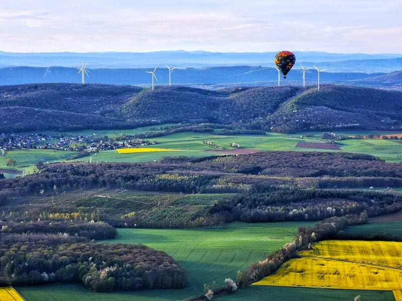 Montgolfière colorée survolant une campagne vallonnée avec champs verts et jaunes, éoliennes en arrière-plan sous un ciel clair.