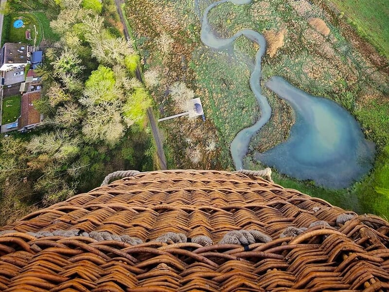 Vue plongeante depuis une nacelle de montgolfière, montrant une rivière et des terres agricoles en contrebas.