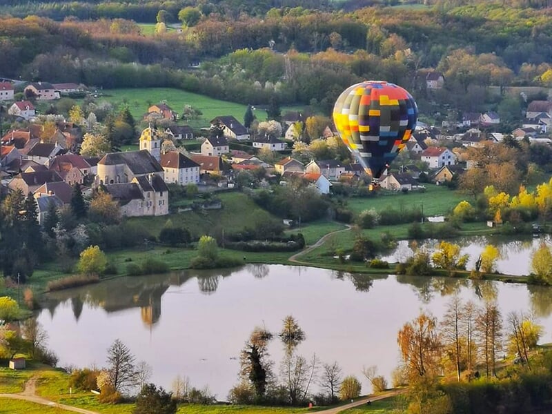 Montgolfière multicolore survolant un lac bordé de maisons et d'arbres dans un village pittoresque.