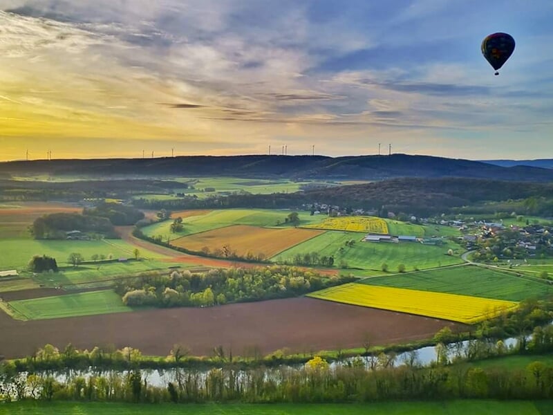Panorama de campagne avec des champs colorés, une rivière et une montgolfière dans le ciel au crépuscule.
