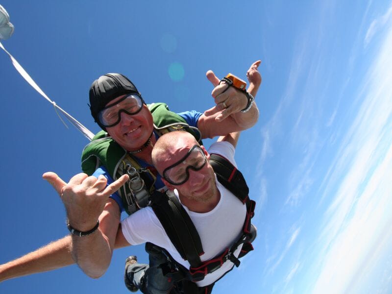 Deux hommes en chute libre pendant un saut en parachute tandem, souriants et faisant des signes de la main, sous un ciel bleu dégagé