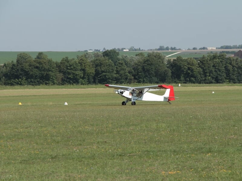 Petit avion blanc et rouge au roulage sur une piste en herbe, entouré d'un paysage verdoyant avec des arbres et des collines en arrière-plan.