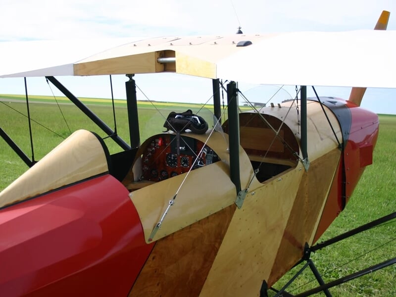 Vue latérale d'un petit avion biplan rouge et beige, posé sur un terrain herbeux avec ciel clair.