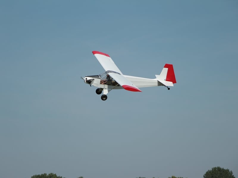 Avion léger blanc avec des extrémités d'ailes et une dérive rouges en plein vol sous un ciel dégagé. L'appareil présente un train d'atterrissage fixe et un cockpit ouvert.