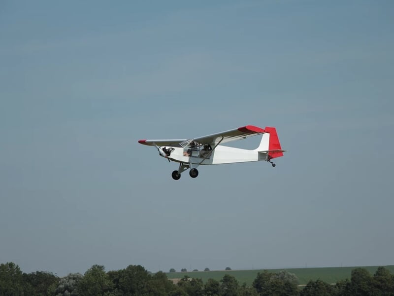 Petit avion blanc et rouge en vol au-dessus d'un paysage avec arbres et ciel bleu clair.