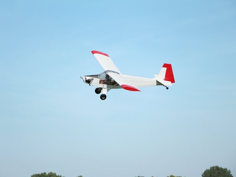 Avion ultra-léger en vol, ailes blanches avec extrémités rouges, arrière rouge, ciel bleu dégagé en arrière-plan.