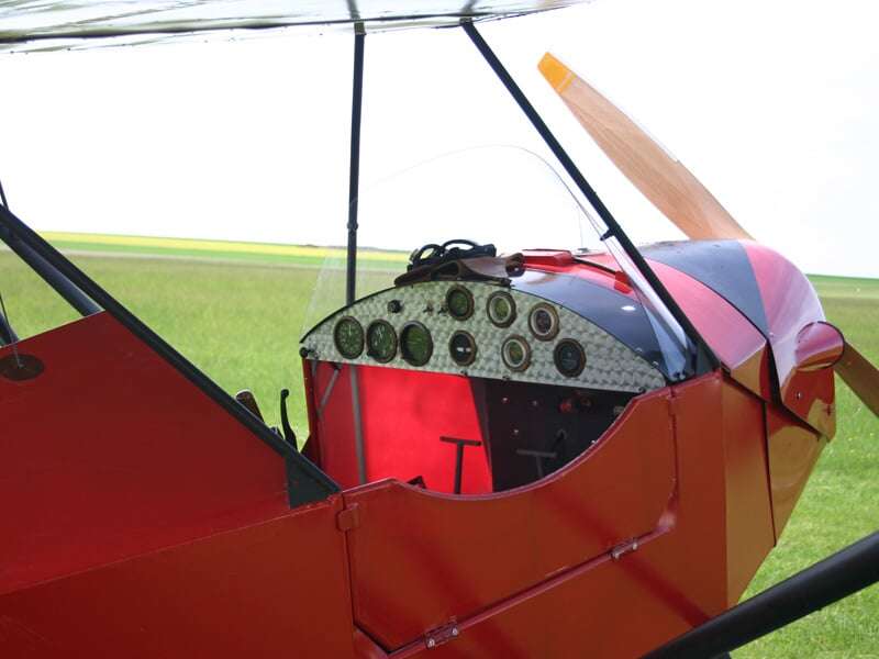 Cockpit d'un petit avion rouge avec tableau de bord visible, posé sur un champ vert sous un ciel clair.