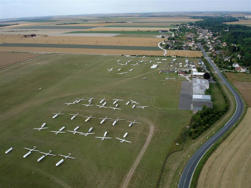 Vue aérienne d’un aérodrome avec planeurs alignés sur une pelouse, bâtiments techniques, routes et champs agricoles alentour.