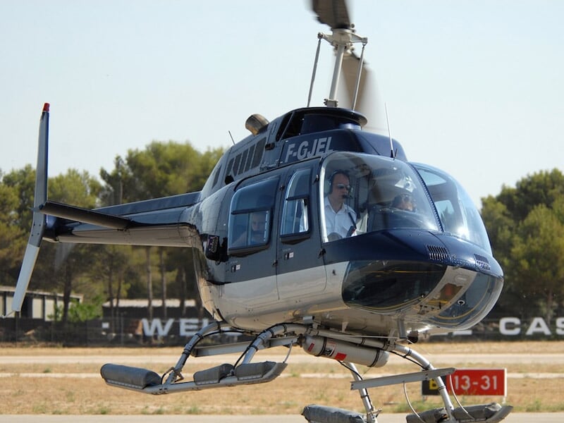 Hélicoptère en vol stationnaire au-dessus d'une piste d'aéroport avec un pilote visible à bord.