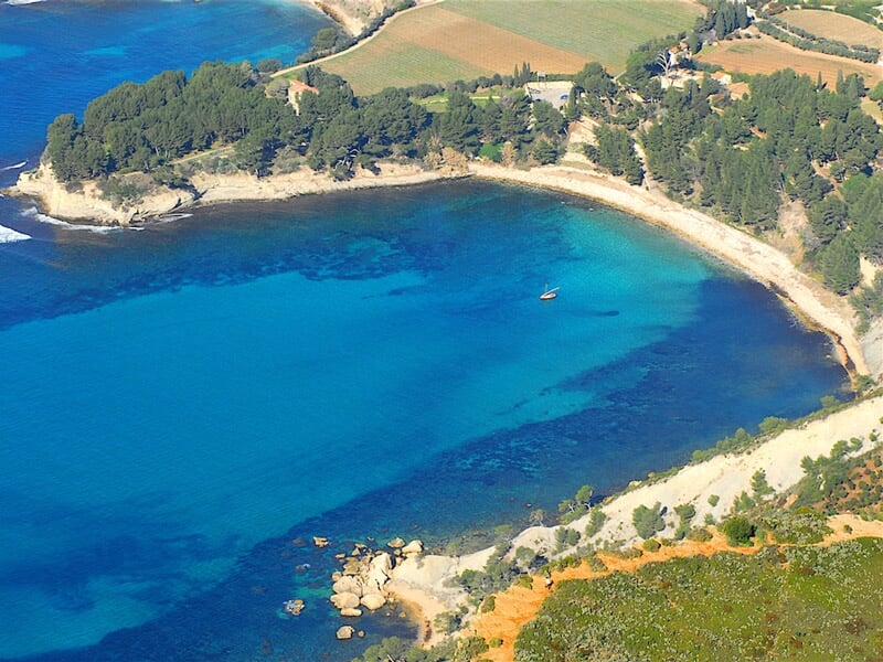 Vue aérienne d'une plage de sable fin entourée de végétation et d'eau turquoise claire sous un ciel ensoleillé.